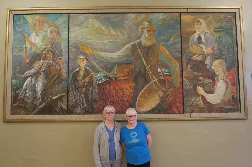 Sisters Dace Ķezbers (right) and Mārīte Plūme, two board members of the Chicago Latvian Association. They're standing in front of a painting titled “Tauta pie jūras,” or “People by the Sea.” The association commissioned prominent Latvian painter Augusts Annusin in 1965 to create it.
